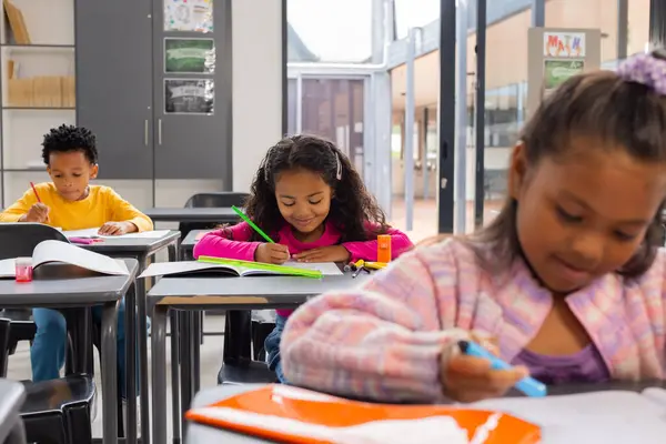 アフリカ系アメリカ人の少年と2人の女性は教室で学校の仕事に集中した 黄色いシャツの男の子はノートブックで書いていますが カラフルな衣装の女の子はマーカーで描きます — ストック写真