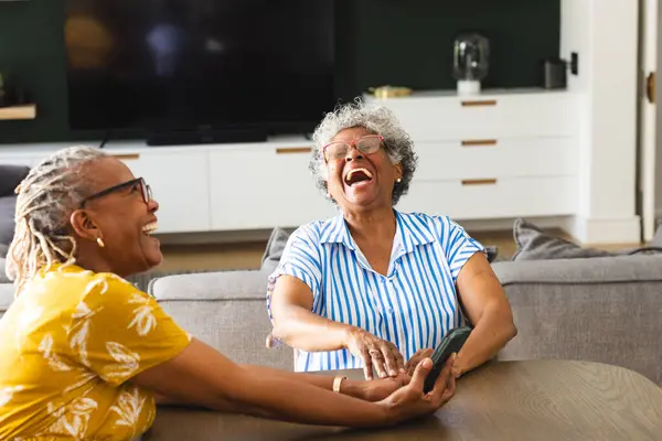 Senior African American woman and senior biracial woman share a laugh together on a couch at home. They are in a cozy living room, bonding over a joyful conversation.