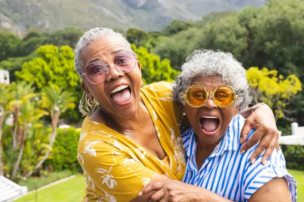 Senior African American woman and senior biracial woman share a joyful embrace outdoors. Both are laughing heartily, showcasing a close bond amidst a vibrant garden setting.