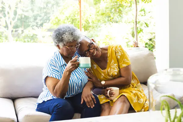 Senior African American woman and senior biracial woman share a cozy moment with coffee at home. They are seated closely on a sofa, exuding warmth and companionship in a bright living room.