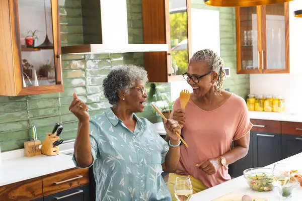 Senior African American woman and senior biracial woman share a moment in a kitchen. They are engaged in a lively conversation, surrounded by cooking utensils and fresh ingredients.