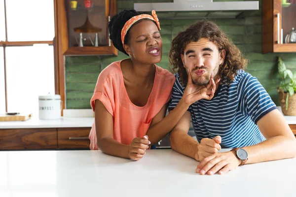 Diverse couple young African American woman playfully pinches the cheek of a young Caucasian man. Both are casually dressed, sharing a light-hearted moment in a bright kitchen setting.