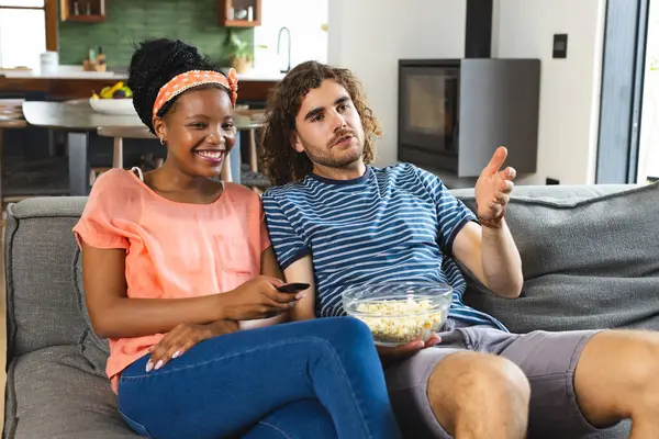 多样化的一对年轻的非洲裔美国女人和白人男人一起在沙发上放松 一起看电视 她笑着戴着头巾 他在谈话中打手势 中间夹着一碗爆米花 免版税图库照片