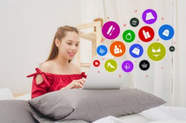 dizüstü bilgisayar kullanarak ve grafik ürün simgesini diyagramı, sosyal ağ teknolojisi, Internet, ödeme ve online alışveriş kavramı ile yatak oda evde oturan kırmızı elbiseli Genç Asyalı güzel kadın