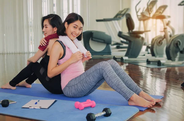 two happy asian young woman slim body with towel sitting with back to each other on yoga mat in fitness gym at morning, bodybuilder, healthy lifestyle, exercise, workout, sport training concept
