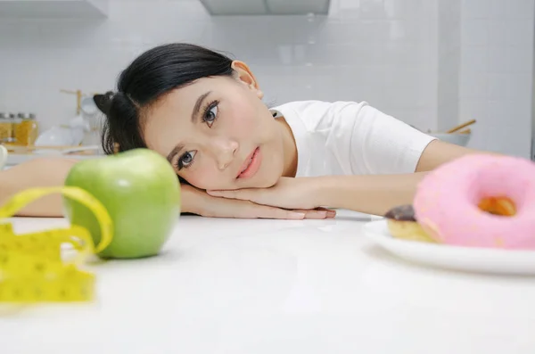 portrait of confused young asian woman choosing between fresh green apple and donut on table in kitchen, healthy food, clean eating, dieting, unhealthy food, good health care and weigh loss concept