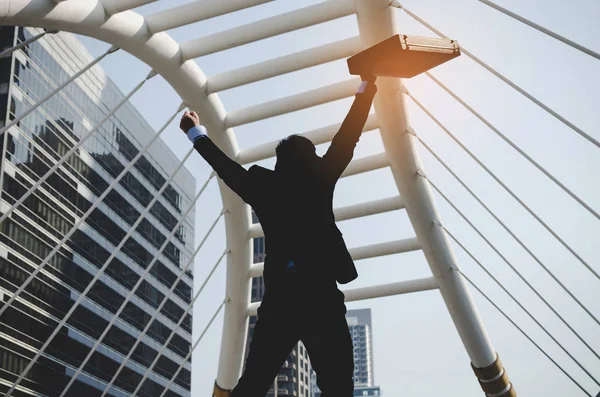 back view of happy young investor business man holding briefcase and raising his fist in the air with modern building in city background, celebrating,  investment and company success concept