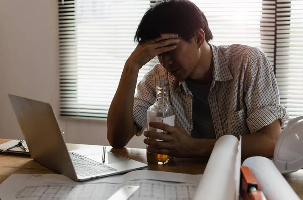 young asian engineer, architect, contractor thinking and worrying about new project contract plan with laptop and bottle of alcohol on desk in office center at construction site, alcoholism concept