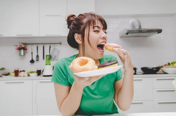 beautiful young asian woman slim body in green shirt enjoying eating delicious glazed donuts and sitting in kitchen interior in house, unhealthy, lifestyle, good healthy, diet food and drink concept