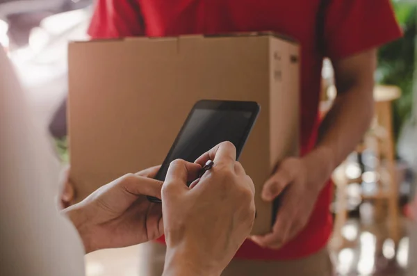 home delivery service man in red uniform and young woman customer appending signature in digital mobile phone receiving parcel post box from courier at home, express delivery, online shopping concept