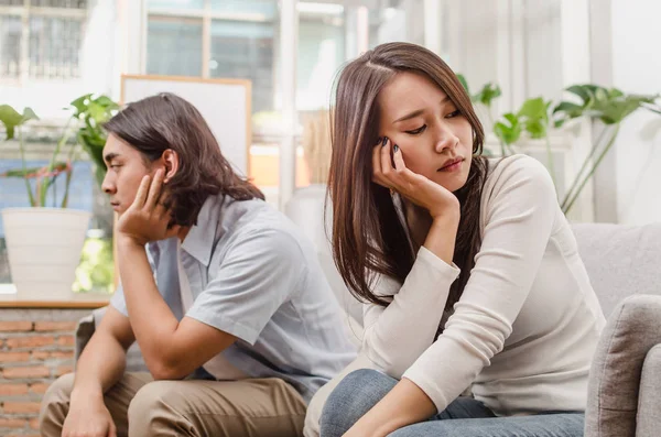 sadness young asian wife having quarrel and sitting on sofa after fight with husband behind her in house interior together, upset couple, love, divorce couple, family issues and relationship concept