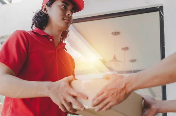 home delivery service handsome man in red uniform handing parcel boxes to recipient and young man customer accepting parcel post box from courier at home, express delivery and online shopping concept