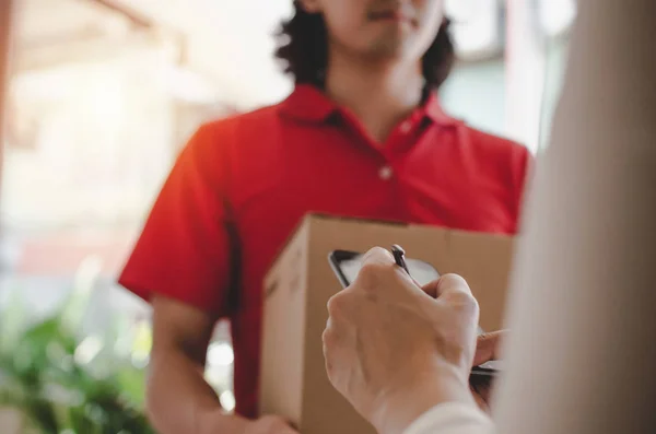 young woman customer appending signature in digital mobile phone receiving parcel post box from courier with home delivery service man in red uniform at home, express delivery, online shopping concept