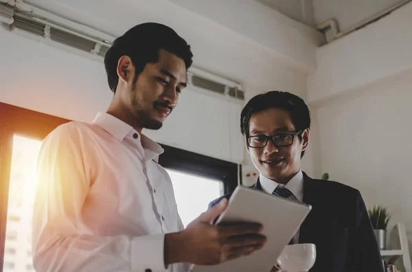 two asian young partner business man investor talking and reading information about financial statistics in mobile tablet together standing in office, finance, investment, digital technology concept