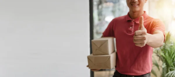 panoramic banner. delivery service man in red uniform showing thumbs up and holding parcel post box ready send to customer at home, express delivery service, online shopping and logistics concept
