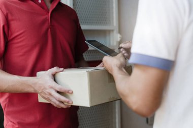 evde kırmızı üniformalı gülen yüz yüze gelen ev teslim servisi ile kuryeden paket posta kutusu alan cep telefonuna dijital imza ekleyen genç adam, ekspres teslimat konsepti