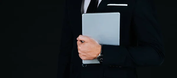 Website banner. young handsome business man in modern black suit holding mobile tablet and standing posture on dark background with copy space, businessman fashion and mobile technology concept