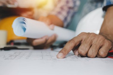 İnşaat şantiyesi, müteahhit, mühendis, mühendislik ve inşaat konseptindeki toplantı odası projesinde çalışma masasının üzerinde plan ve güvenlik kaskı üzerinde çalışan grup mühendisi ve mimarın elleri