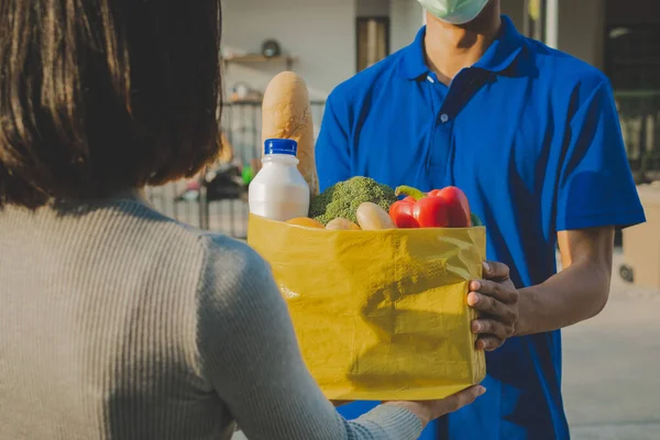 delivery service man with protection face mask in blue uniform holding fresh food set bag for customer at door home, grocery, healthy food, express delivery, food delivery, online shopping concept