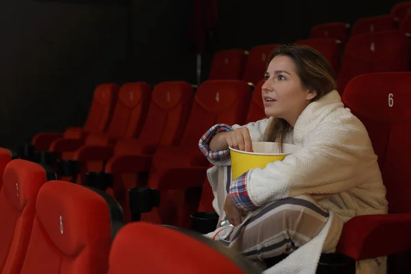 A interested girl in a robe or pajamas sits in the cinema alone watching a movie and eating popcorn