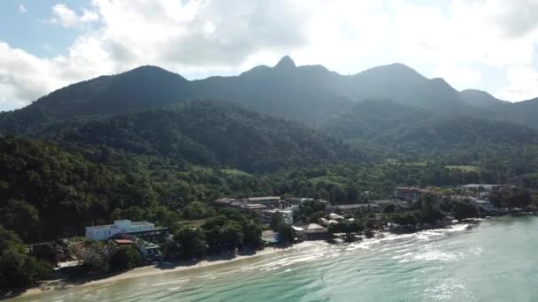 Die grünen Hügel der Insel sind mit Palmen bedeckt, der Himmel ist bewölkt und ein Sonnenstrahl bricht durch. Klares türkisfarbenes Wasser am Strand mit goldenem Sand. Die Leute entspannen sich. Drohne. — Stockvideo