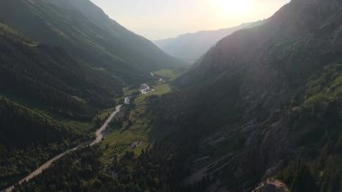 Warm sunlight illuminates a picturesque mountain valley, revealing a winding river flowing through lush greenery and a road snaking alongside it