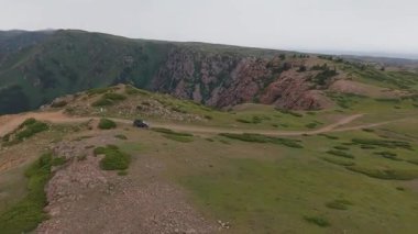 Arazi aracı manzaralı bir dağ sırtı boyunca virajlı toprak bir yolda ilerliyor, aşağıdaki vadiyi nefes kesen manzaralar sunuyor ve uzaktaki karlı zirveler.