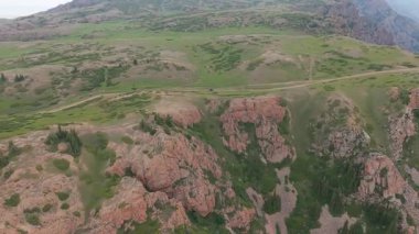 Görkemli dağ arazisi engebeli kayalık araziyi gözler önüne seriyor, yoğun olarak canlı yeşil bitki örtüsüyle kaplı, dramatik bulutlu ufuk çizgisinin altında geniş bir vahşi doğa manzarası var.
