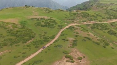 Yeşil çayırlar ve çalılar yuvarlanan tepeleri kaplıyor. Siyah bir SUV virajlı toprak bir yolda giderken, bir dağ sırası uzaklıktan görülebiliyor.