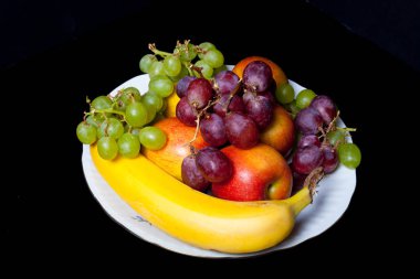 Siyah bir zemin üzerine beyaz seramik tabakta meyvelerin farklı türleri.