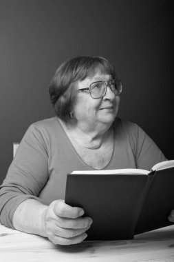 Gözlüklü yaşlı bir kadın bir masada oturur ve bir kitap okur. Tonda.