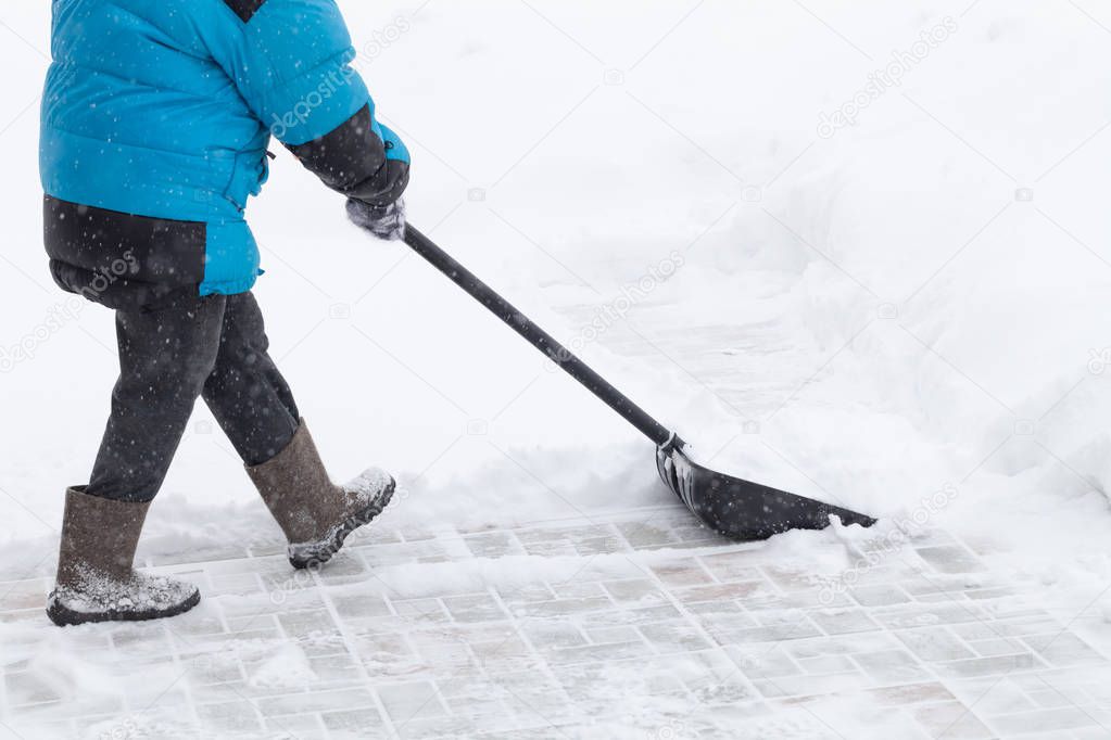 Old woman in warm blue jacket clears a snowdrifts with a snow sh