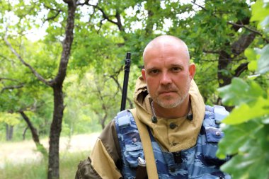 Ava. 35-40 yaşında bir adam, ormanda bir ateşli silah ile ciddi bir yüz ile askeri bir avcı duruyor.