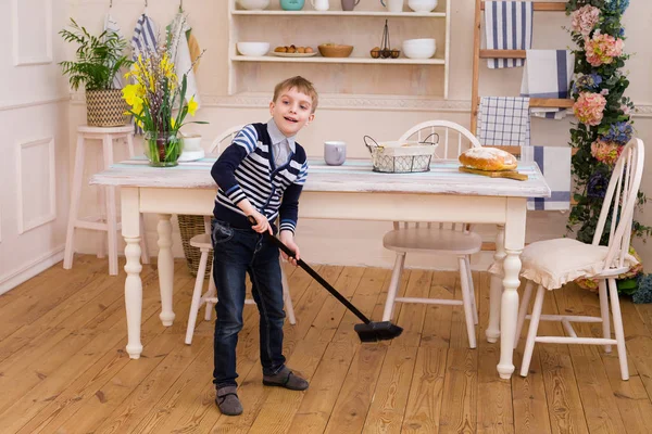 Blond boy sweeping the floor in the kitchen. Pretty boy helps parents with housework. Child cleaning room with broom. Child cleaning kitchen. Housekeeping concept.