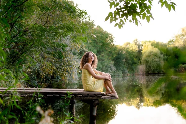 卷发的年轻女子 穿着黄色的衣服坐在湖边 夏日休闲 幸福的理念 — 图库照片