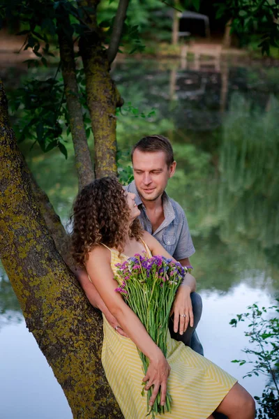 Couple in love, happy woman with flowers, summer time. Green trees outdoor, family couple near lake, love and romantic. Concept of relationship, happiness, togetherness.