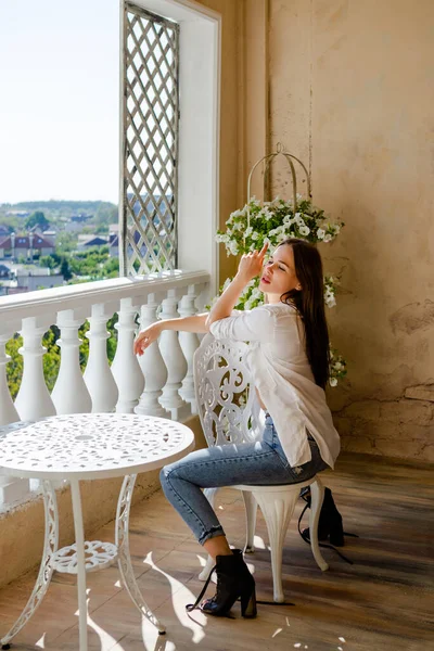 Masanın yanındaki terasta oturan çekici bir kadın. Haftasonu konsepti ve rahatlama. Mavi kot ve beyaz gömlek giyen siyah saçlı kadın..