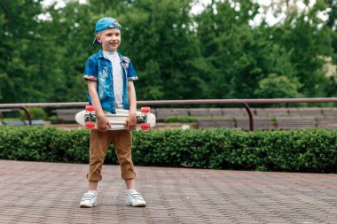 Bir küçük şehir çocuğu ve bir kaykay. Genç bir adam parkta duran ve bir kaykay tutarak. City Style. Şehir çocukları. Bir çocuk bir kaykay binmek öğrenir