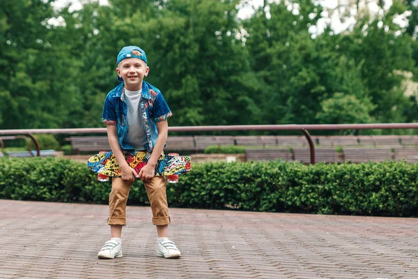 A small city boy and a skateboard. A young guy is standing in the park and holding a skateboard. City Style. City children. A child learns to ride a skateboard