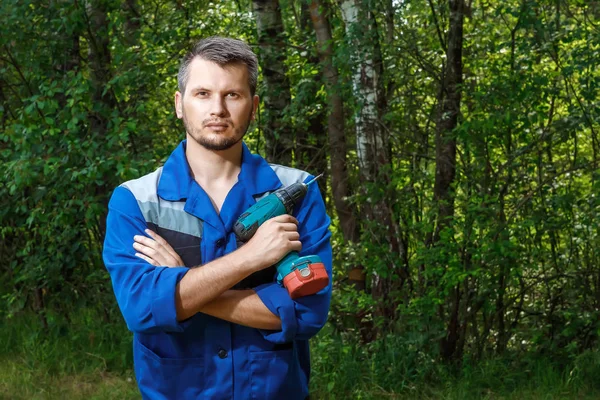 A man is a builder in a robe with a screwdriver in his hands, a coveralls against the background of a green forest. Isolate, copy space.