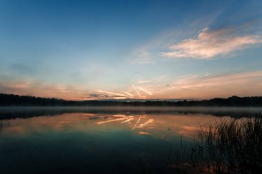 Güzel, kırmızı şafak göl kenarında. Sis sayesinde güneş ışınları. Göl üzerinde mavi gökyüzü, sabah geliyor, orman suya yansıyan.
