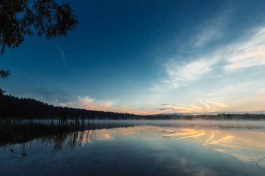 Güzel, kırmızı şafak göl kenarında. Sis sayesinde güneş ışınları. Göl üzerinde mavi gökyüzü, sabah geliyor, orman suya yansıyan.