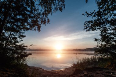 Güzel, kırmızı şafak göl kenarında. Sis sayesinde güneş ışınları. Göl üzerinde mavi gökyüzü, sabah geliyor, orman suya yansıyan.