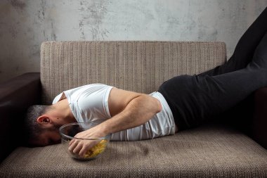 Gömlekli adam kanepede yatıyor, cips yiyor ve spor kanalını izliyor. Tembellik kavramı, hayal kırıklığı, erteleme, evde kişi.
