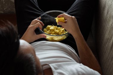 Gömlekli adam kanepede yatıyor, cips yiyor ve spor kanalını izliyor. Tembellik kavramı, hayal kırıklığı, erteleme, evde kişi.