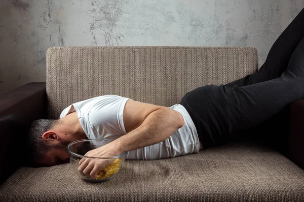 The guy in the shirt is lying on the couch, eating chips and watching a sports channel. The concept of laziness, frustration, procrastination, the person at home.