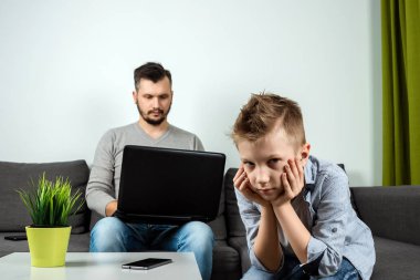 Babası evde arka planda çalışırken üzgün bir çocuk kameraya benziyor. Freelancing, kavramı zaman birlikte, yalnız çocuk, evde, iş kopya alanı.