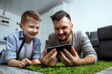 Mutlu aile, baba ve oğul birlikte yerde, gülümseyerek, bir smartphone bir şey izlerken yatıyor. Aile coşku, kavramı zaman geçirdiyseniz, alanı Kopyala.
