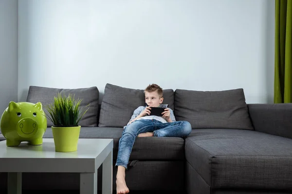 A boy sits on a sofa in the living room and plays with a smartphone. Boy texting on the phone. The concept of harming modern gadgets.