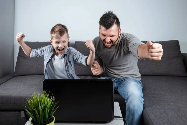 Father and son watching football in a laptop at home. Emotional man and boy, cheering favorite team. The concept of family enthusiasm, emotions, family time, copy space.